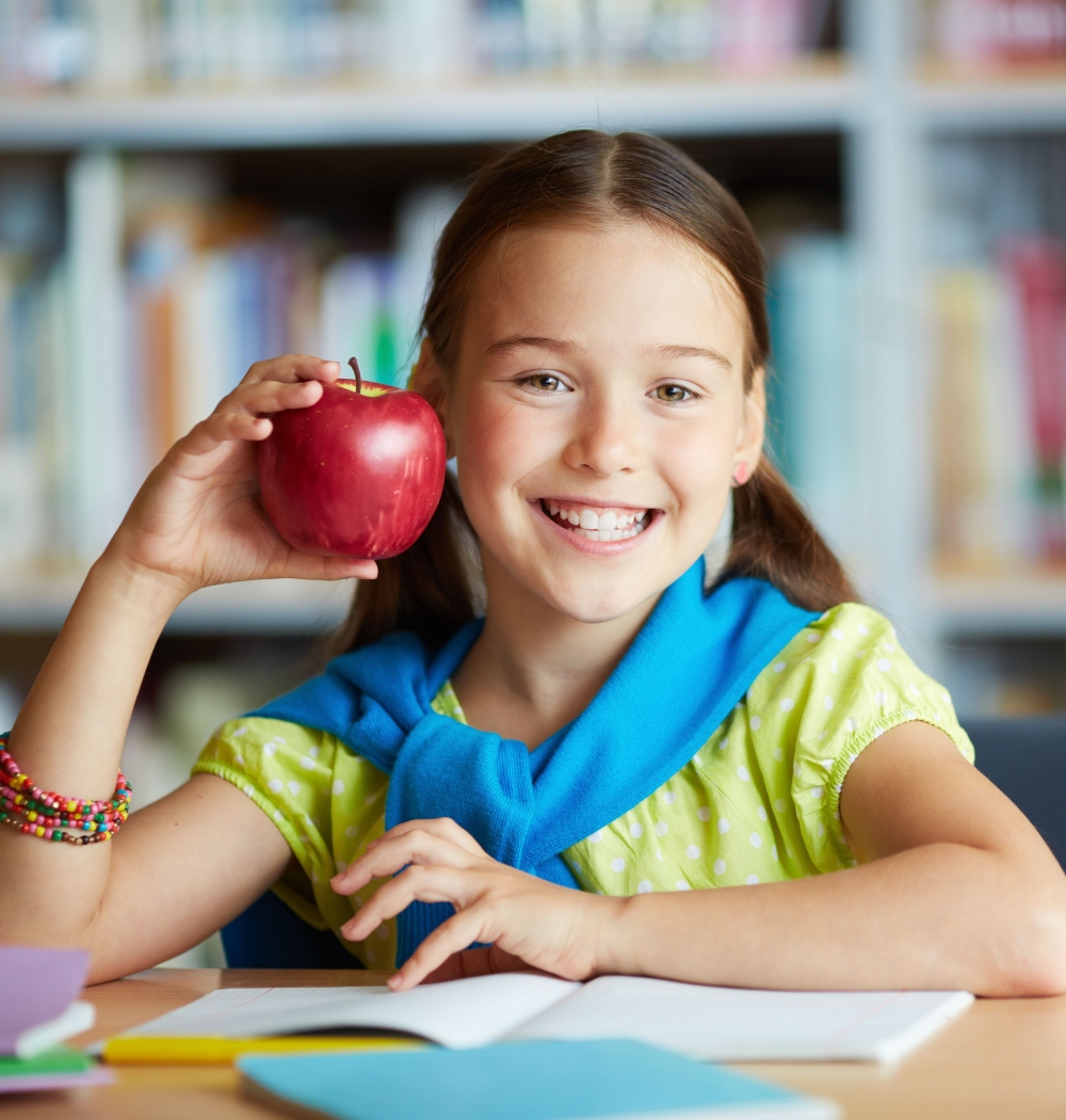 Portrait of happy schoolgirl with big red apple looking at camera in library